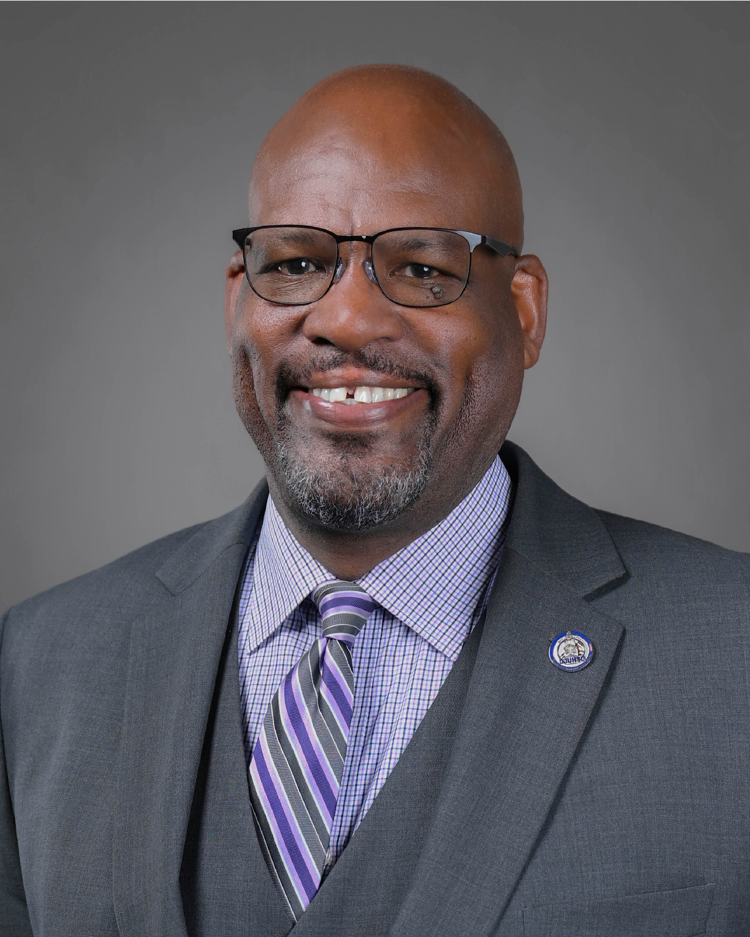 Smiling man with goatee and glass wearing a grey suit with a purple and grey tie.